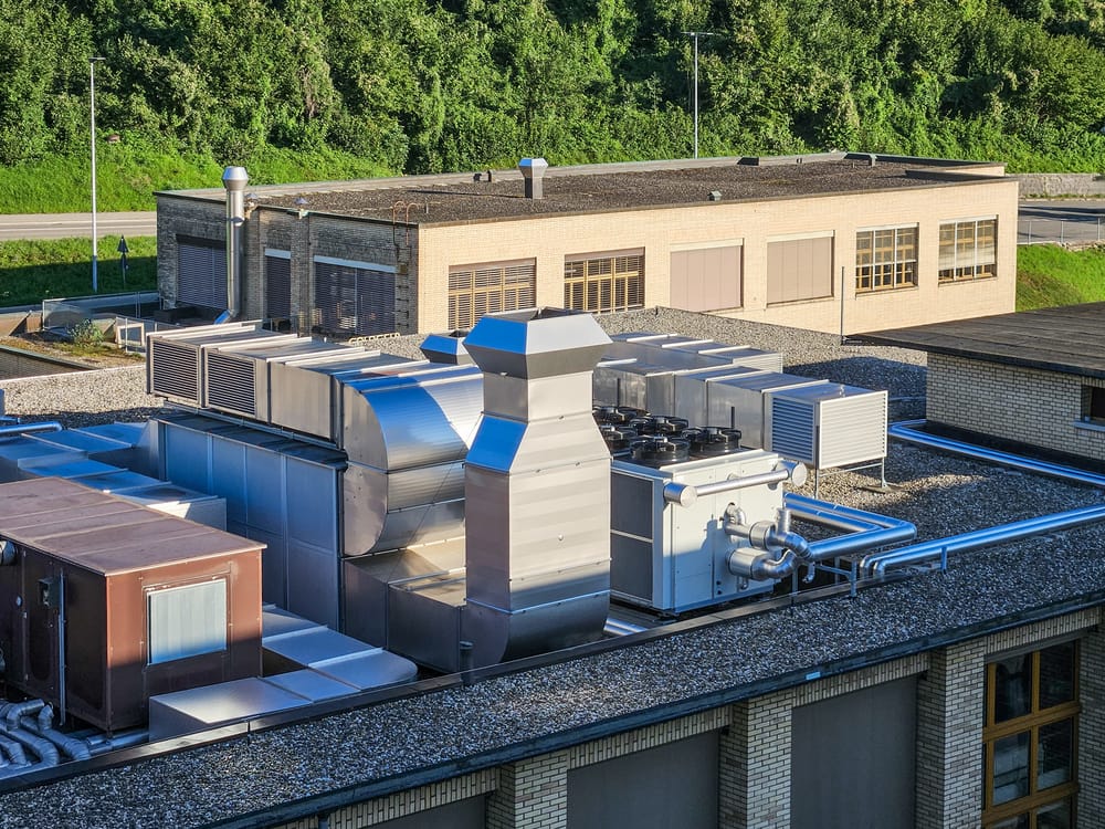 Rooftop view of a commercial building with HVAC systems and ventilation ducts. A small administrative building and trees are visible in the background.