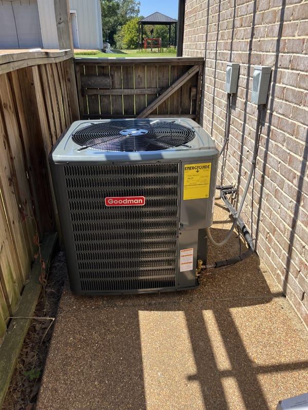 Outdoor Goodman air conditioning unit installed on a paved pathway near a brick wall and wooden fence, with cables connected to wall-mounted electric boxes.
