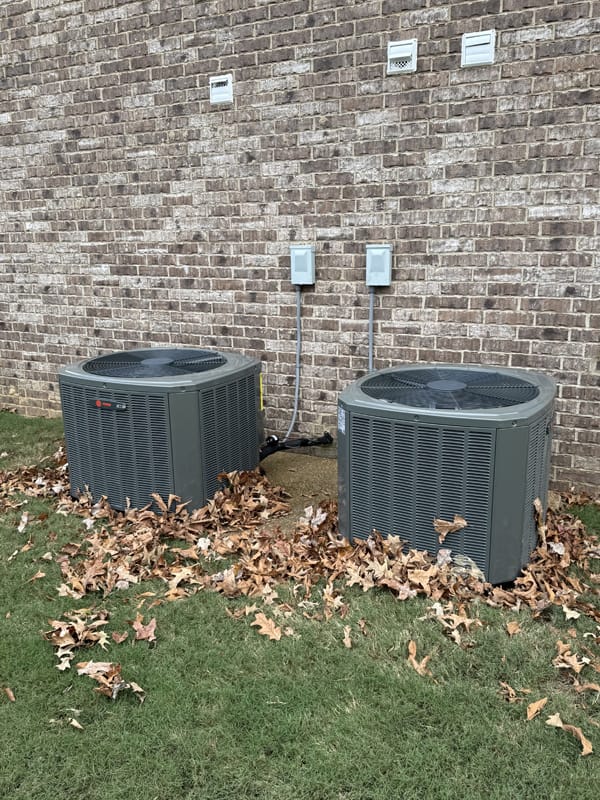 Two outdoor air conditioning units next to a brick wall, surrounded by fallen leaves on the grass.